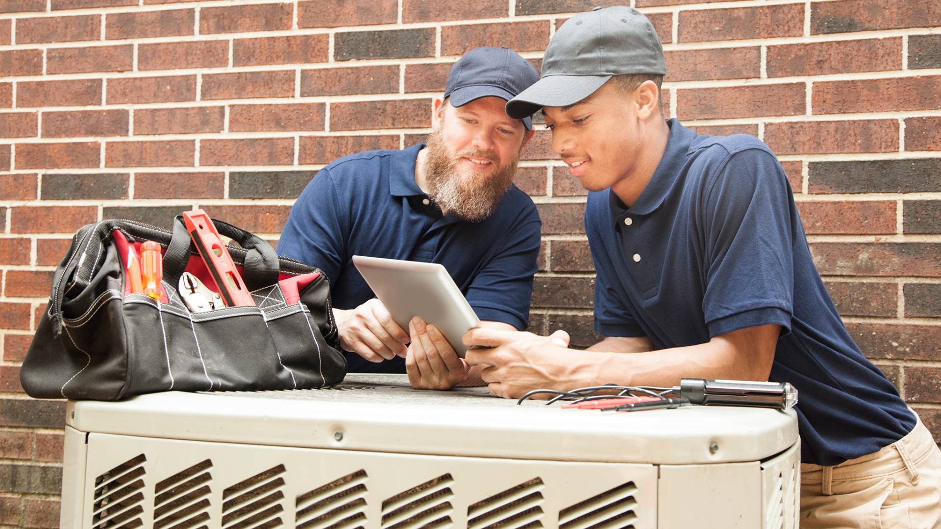 two HVAC techs looking at a tablet and resting on an HVAC unit